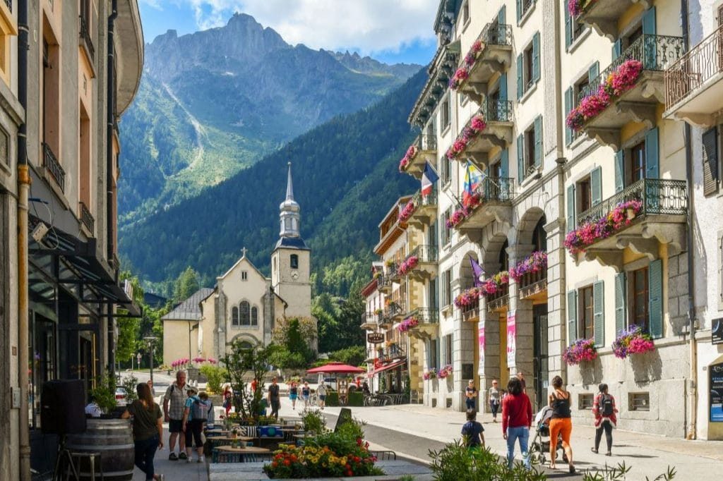 Classe-Affaires VTC Chamonix Mont-Blanc Rue animée avec bâtiments fleuris et une église au fond, entourée de montagnes.