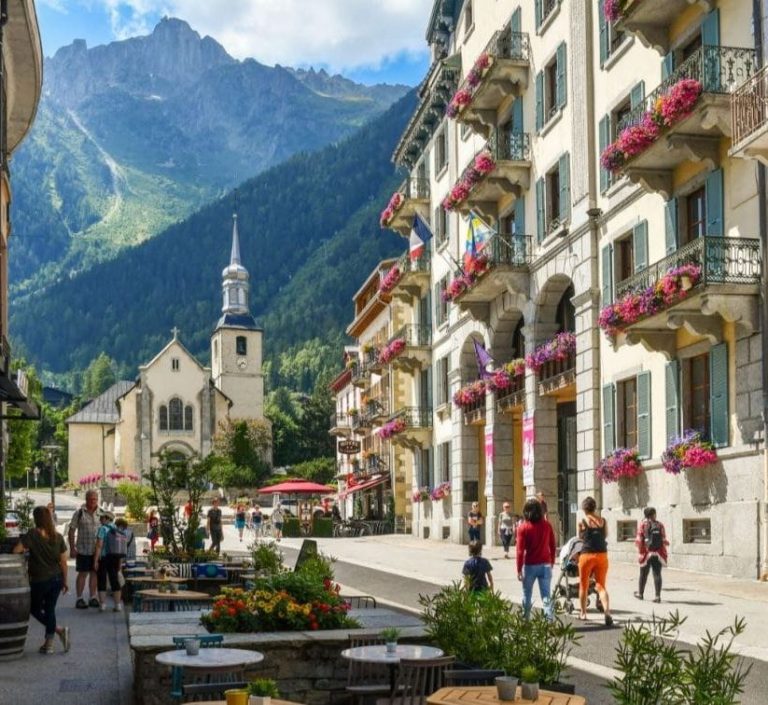Classe-Affaires VTC Chamonix Mont-Blanc Rue animée à Chamonix Mont-Blanc, avec des montagnes en arrière-plan et une église.