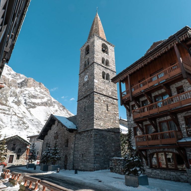 Classe-Affaires VTC Chamonix Mont-Blanc Val d'Isère Église en pierre avec un clocher à Val d'Isère, entourée de montagnes enneigées et de chalets.